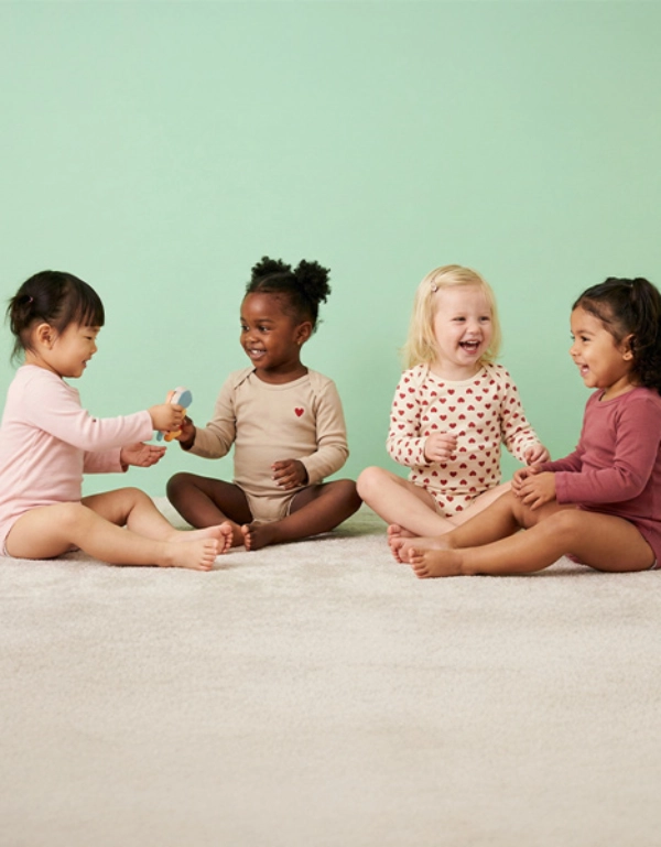 Eine Gruppe von vier Kleinkindern sitzt auf einem hellen Boden und beschäftigt sich gemeinsam mit einem Spielzeug vor einem einfarbigen pastellgrünen Hintergrund.