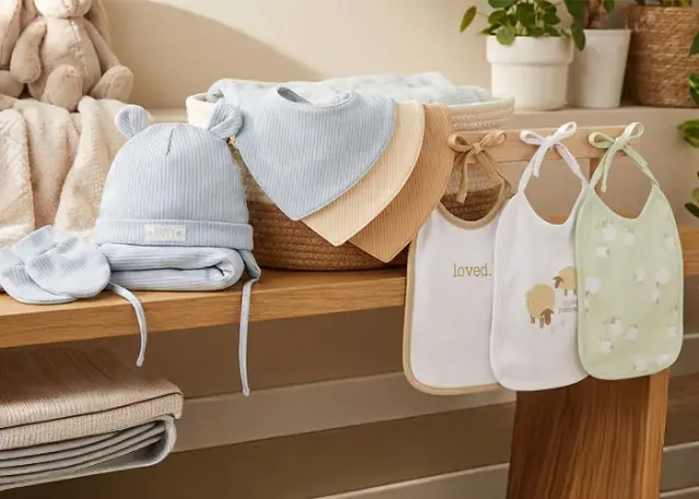 Eine gemütliche Präsentation von Baby-Essentials: eine blaue Mütze, weiche Decken und Lätzchen mit niedlichen Motiven, arrangiert auf einem Holzregal. Eine gemütliche Präsentation von Baby-Essentials: eine blaue Mütze, weiche Decken und Lätzchen mit niedlichen Motiven, arrangiert auf einem Holzregal.