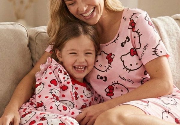 Eine Mutter und ihr Kind kuscheln sich auf einem gemütlichen Sofa, beide tragen passende rosa Hello Kitty-Pyjamas und bunte Socken. Sie sind von einer sanften Einrichtung umgeben.