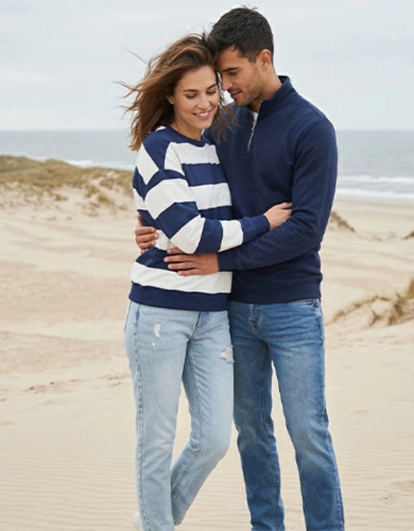 Ein Paar in lässiger Kleidung umarmt sich an einem Sandstrand mit Dünen und Meereswellen unter einem bewölkten Himmel.