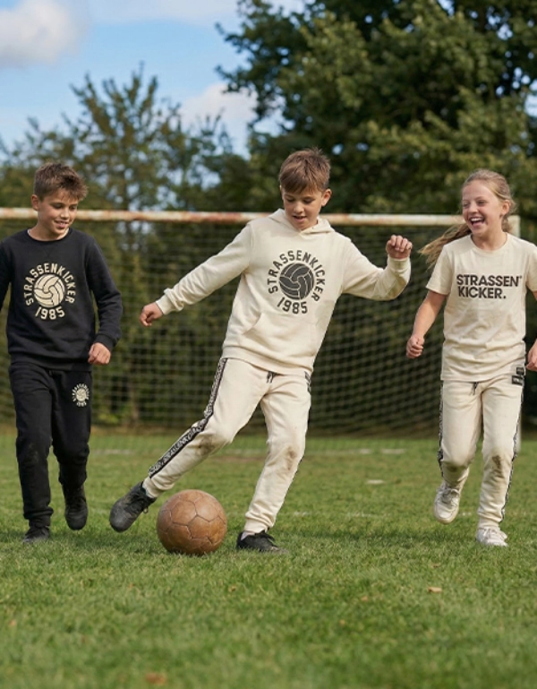 Drei Kinder in lässiger Sportkleidung spielen Fußball auf einer Grasfläche, mit einem Tor im Hintergrund.