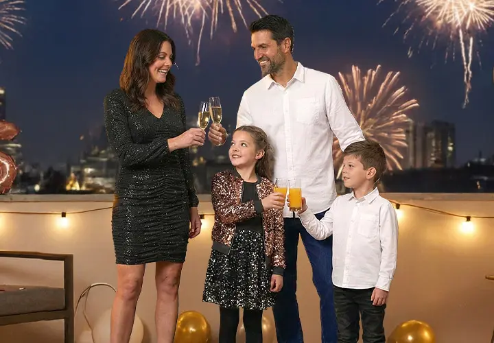 Eine Familie feiert mit Getränken auf einem Balkon, während hinter ihnen ein Feuerwerk den Nachthimmel erhellt.