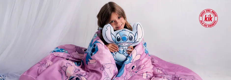 Ein Kind ist in eine rosa Decke mit Stitch-Motiv eingehüllt, hält ein Stitch-Plüschtier und sitzt vor einem weichen weißen Hintergrund.