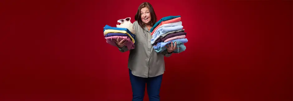 Eine Person in einem grauen Shirt hält große Stapel bunt gefalteter Kleidung, auf denen eine stylische Sonnenbrille und ein Hut liegen.