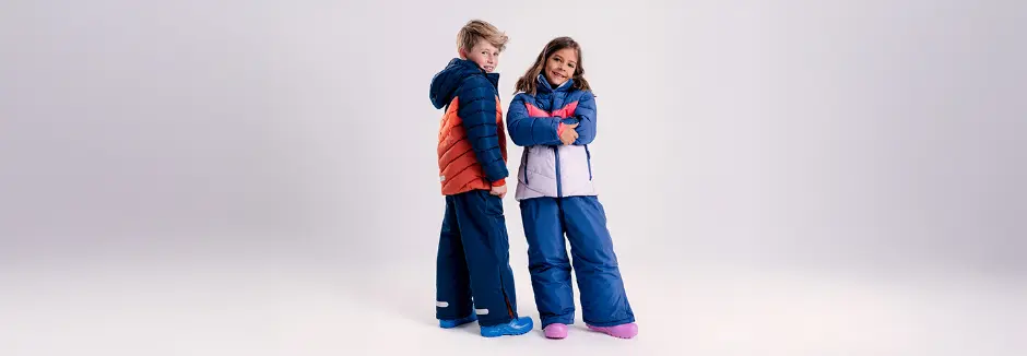 Zwei Kinder stehen Rücken an Rücken und tragen farbenfrohe Winterjacken und Schneehosen vor einem weichen, neutralen Hintergrund.